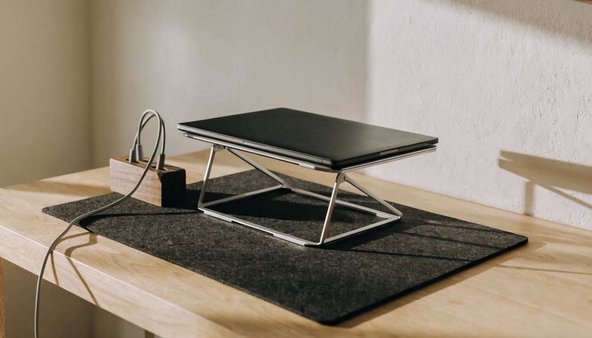 A minimal desk with a cork pad, an aluminium laptop stand and a walnut cable holder in morning light